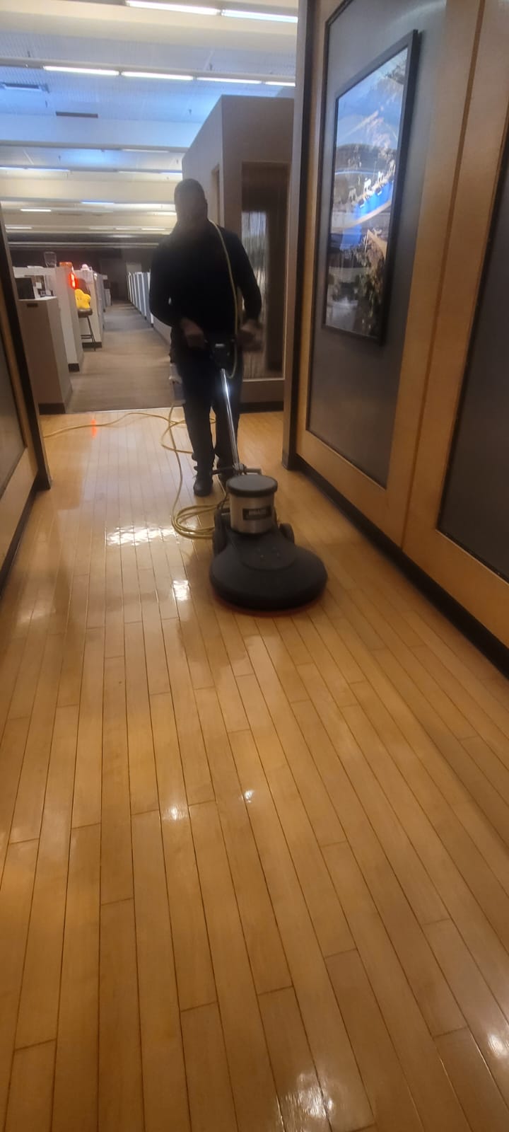 Person polishing a shiny wooden floor with a floor buffer in an office hallway.
