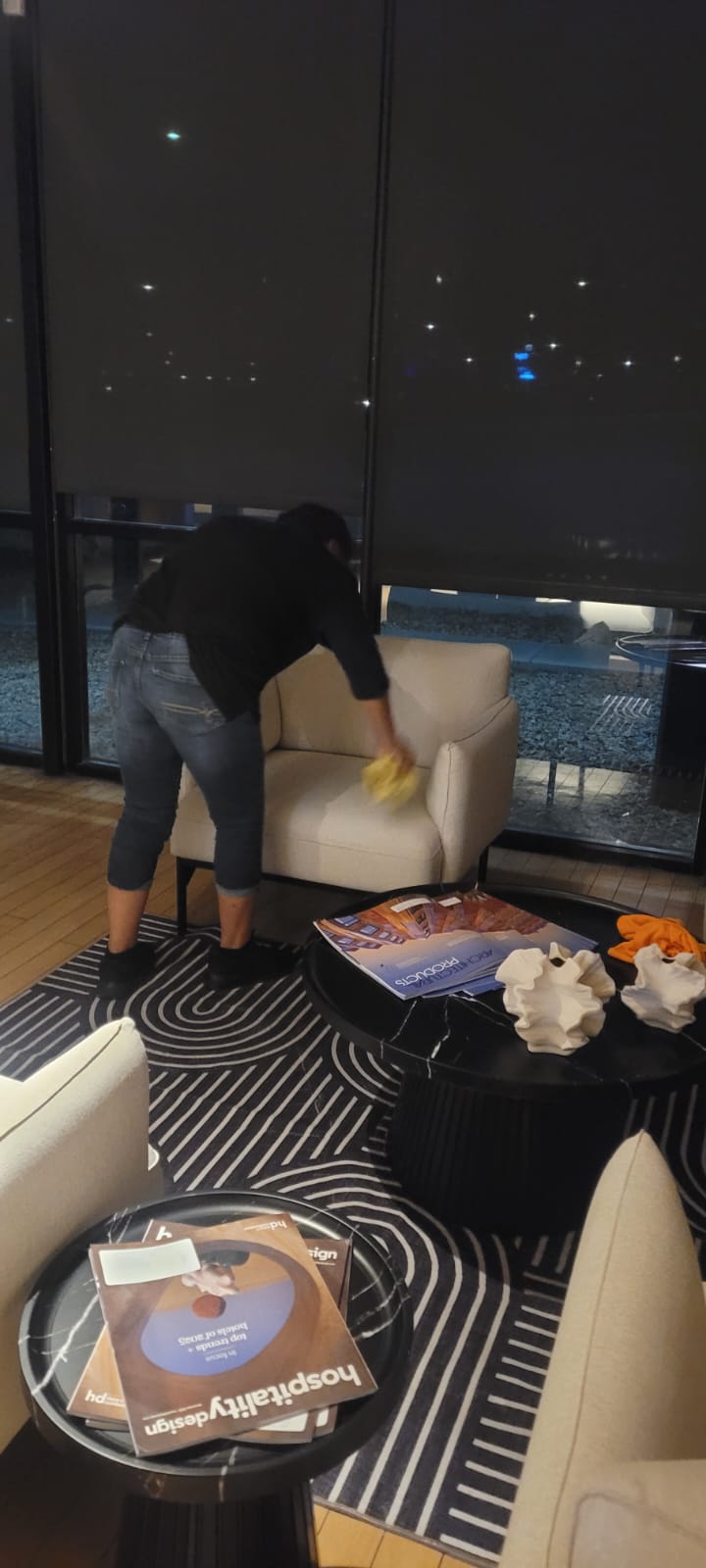 Person cleaning a beige armchair with a yellow cloth in a modern room with black and white patterned rug and coffee tables holding magazines and decorative items.